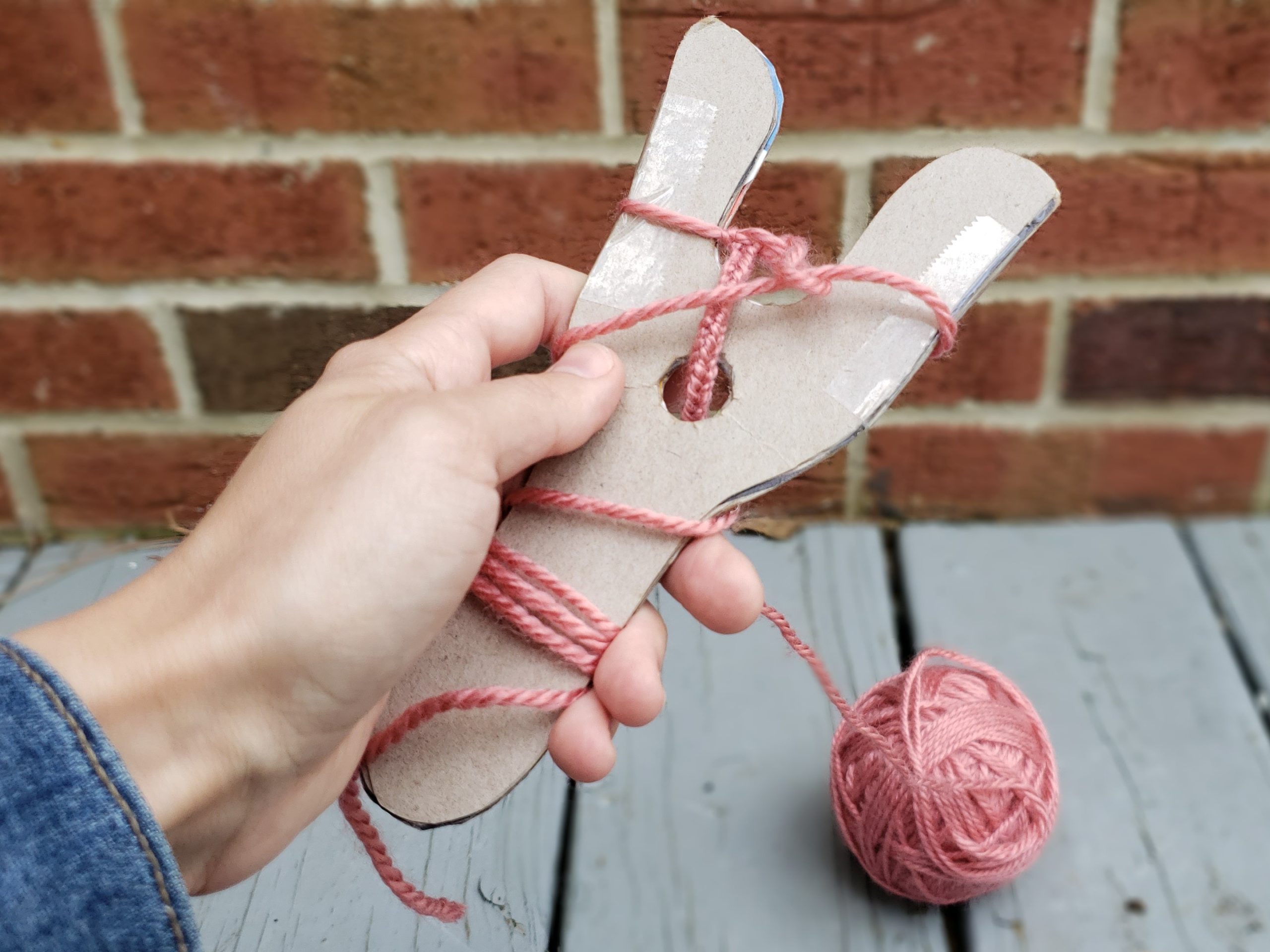 First person point of view of hand holding a sturdy cardboard lucet weaving tool with a work-in-progress wound through it.